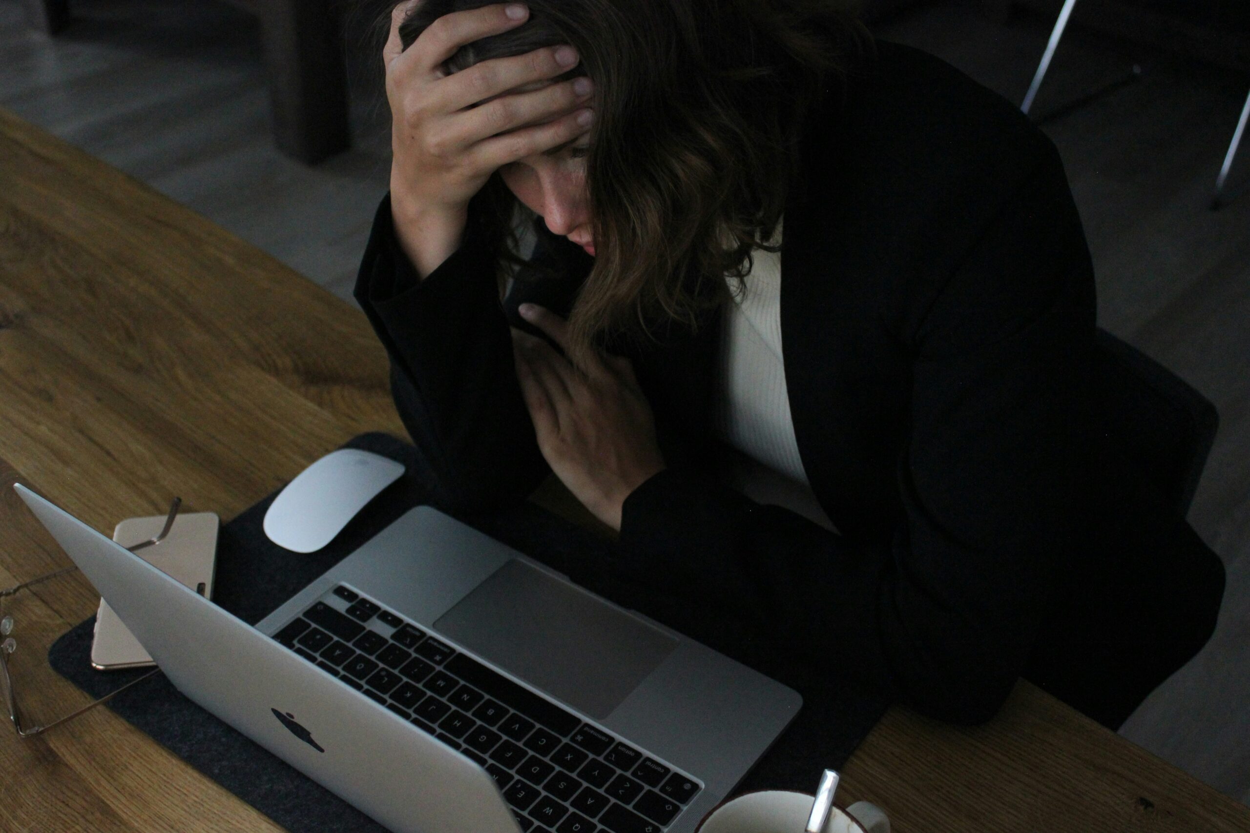 Image of a women at work sitting at a desk, looking at her macbook laptop. She has her head in her hands and looks overwhelmed.